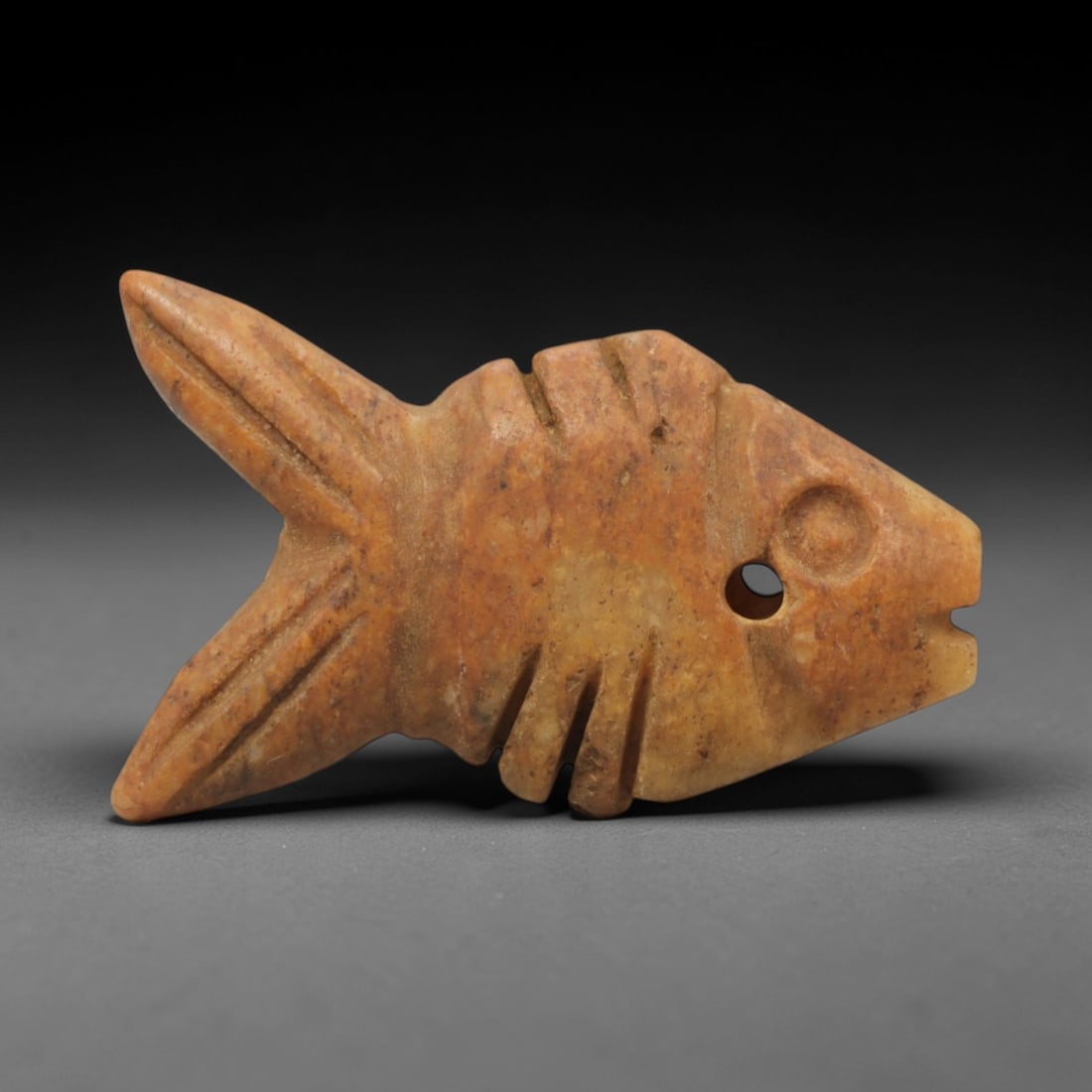 Ancient Stone Fish Amulet - Egyptian Pendant Figurine, 17XR: Carved from a warm ochre-toned stone, this amulet depicts a stylized fish with incised linear details defining its fins, body segments, and a distinct circular eye, with a perforation at the head for