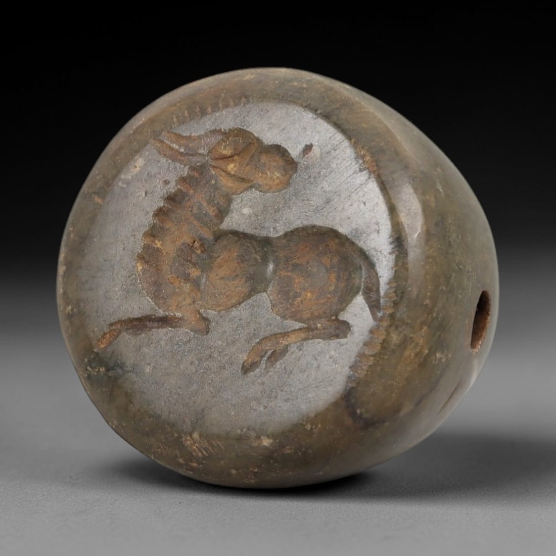 Ancient Steatite Conoid Stamp Seal - Animal Motif, 38AH: Carved from steatite, this ancient conoid stamp seal features a raised relief of a stylized animal motif on the upper surface, with a central perforation for suspension. The stone exhibits a muted gre