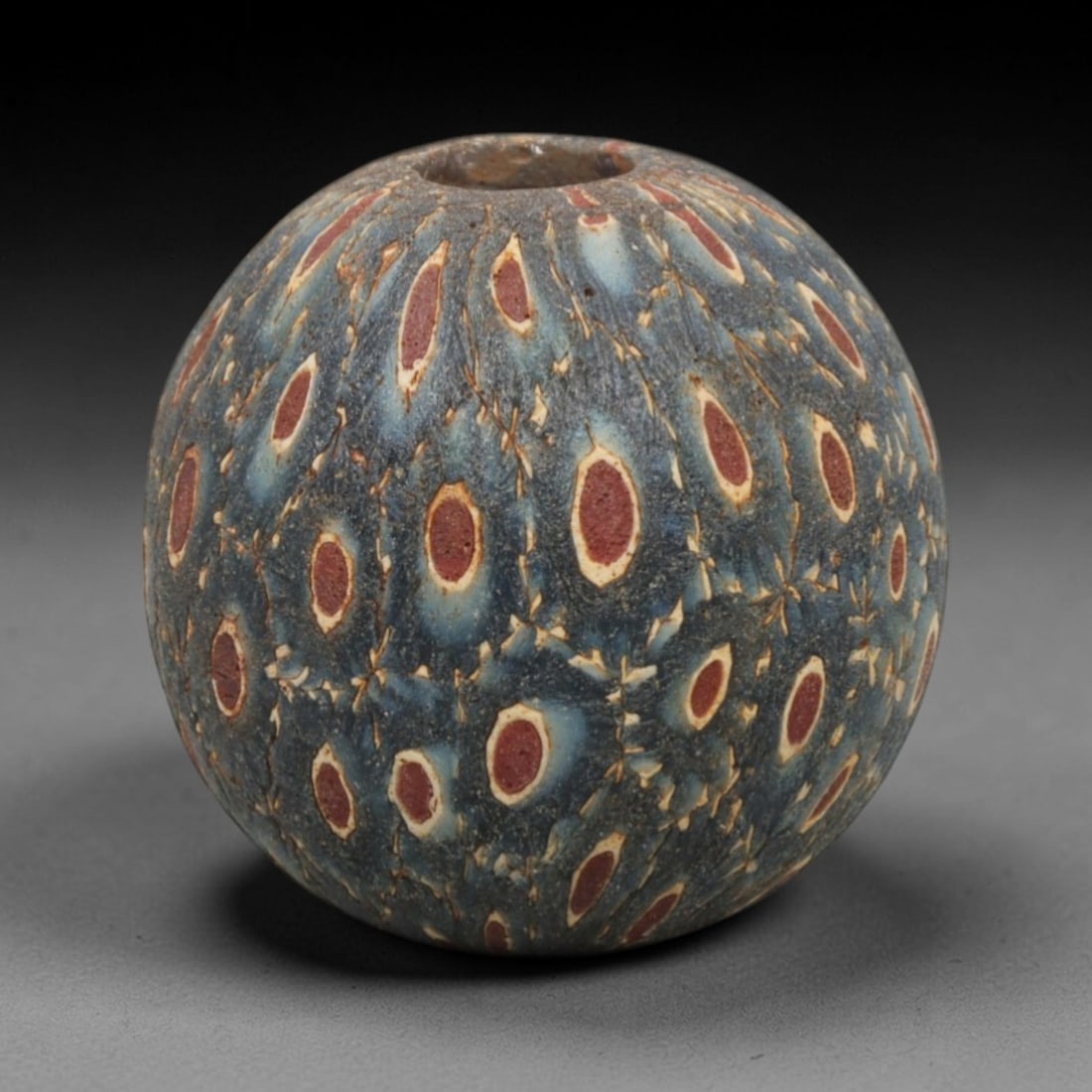 Ancient Glass Bead - Roman Egyptian Peacock Eye Amulet, 85ZN: Crafted from mosaic glass, this spherical bead features a central suspension hole and is decorated with repeating peacock eye motifs in red, white, and blue glass inlay, characteristic of Roman-Egypti