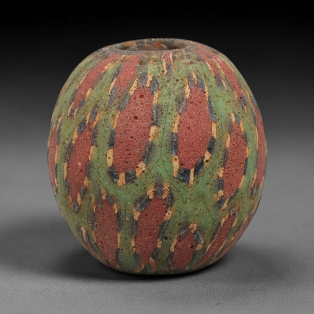 Ancient Islamic Mosaic Glass Bead with Red-Green Pattern, 76ZN: A fine example of an ancient Islamic mosaic glass bead, dating to approximately the 8th-12th centuries CE. This barrel-shaped bead features a striking geometric pattern of deep red oval panels interwo