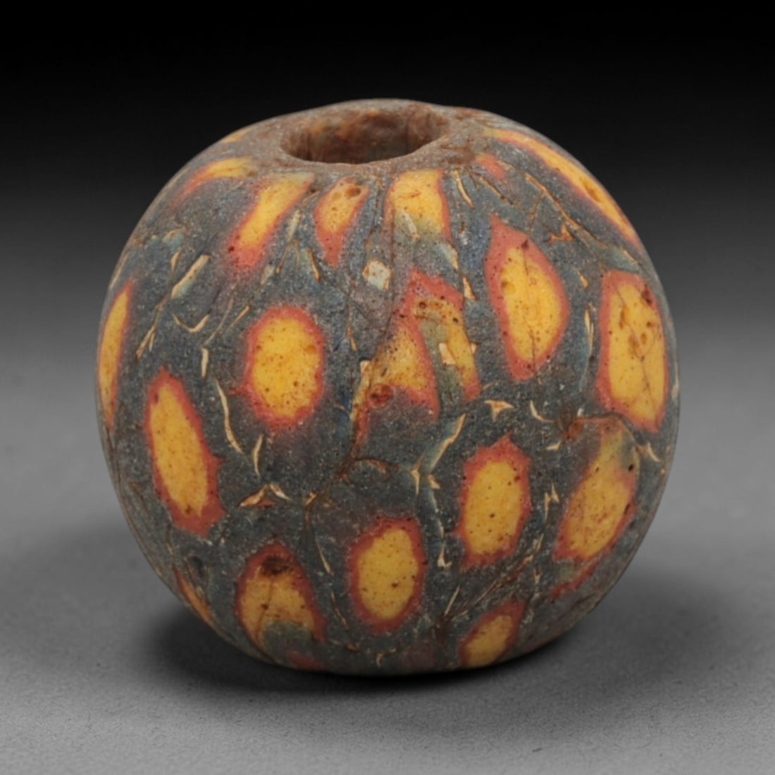 Ancient Glass Bead - Roman Egyptian Mosaic Amulet, 53ZN: Crafted from mosaic glass, this spherical bead features a central suspension hole and is decorated with repeating teardrop-shaped motifs in yellow, red, and blue glass inlay, characteristic of Roman-E