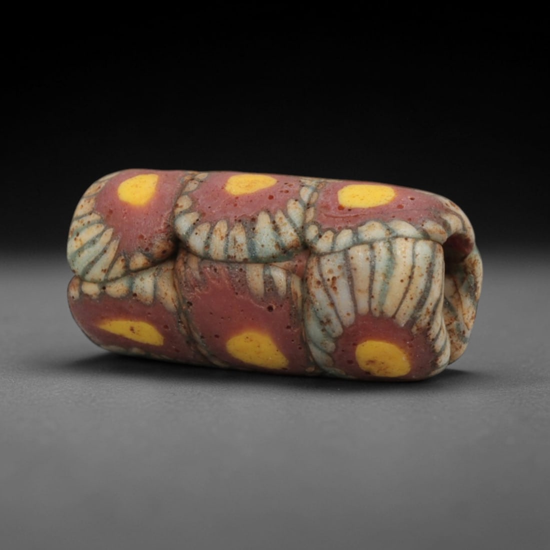Ancient Islamic Floral Mosaic Glass Bead, 61ZN: A rare elongated ancient Islamic mosaic glass bead, dating to the 8th-12th centuries CE. This cylindrical bead showcases a vibrant floral mosaic pattern, with deep red panels adorned with bright yello