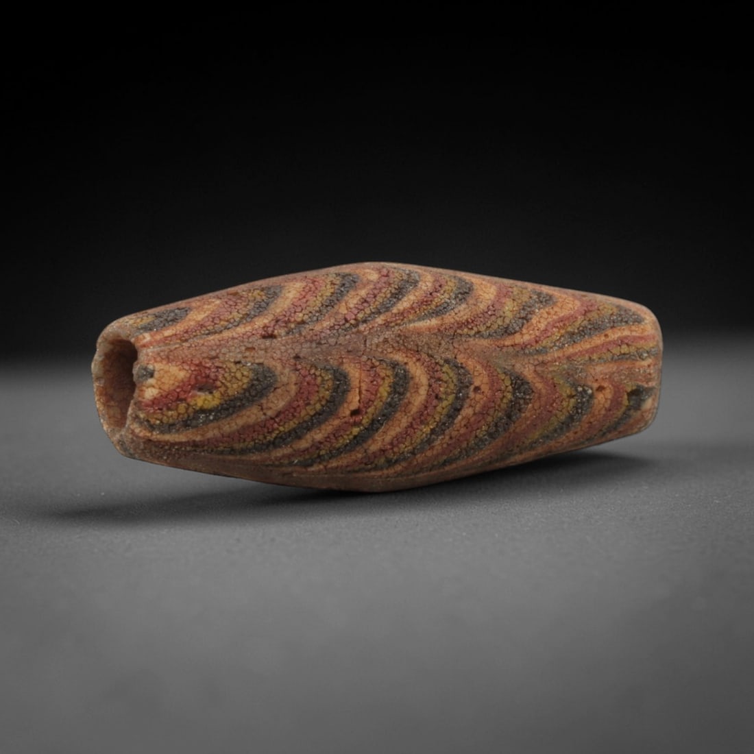 Ancient Islamic Feather-Pattern Mosaic Glass Bead, 60ZN: A rare elongated ancient Islamic mosaic glass bead, dating to the 8th-12th centuries CE. This spindle-shaped bead showcases an intricate feather-like pattern of alternating red, yellow, and dark grey/