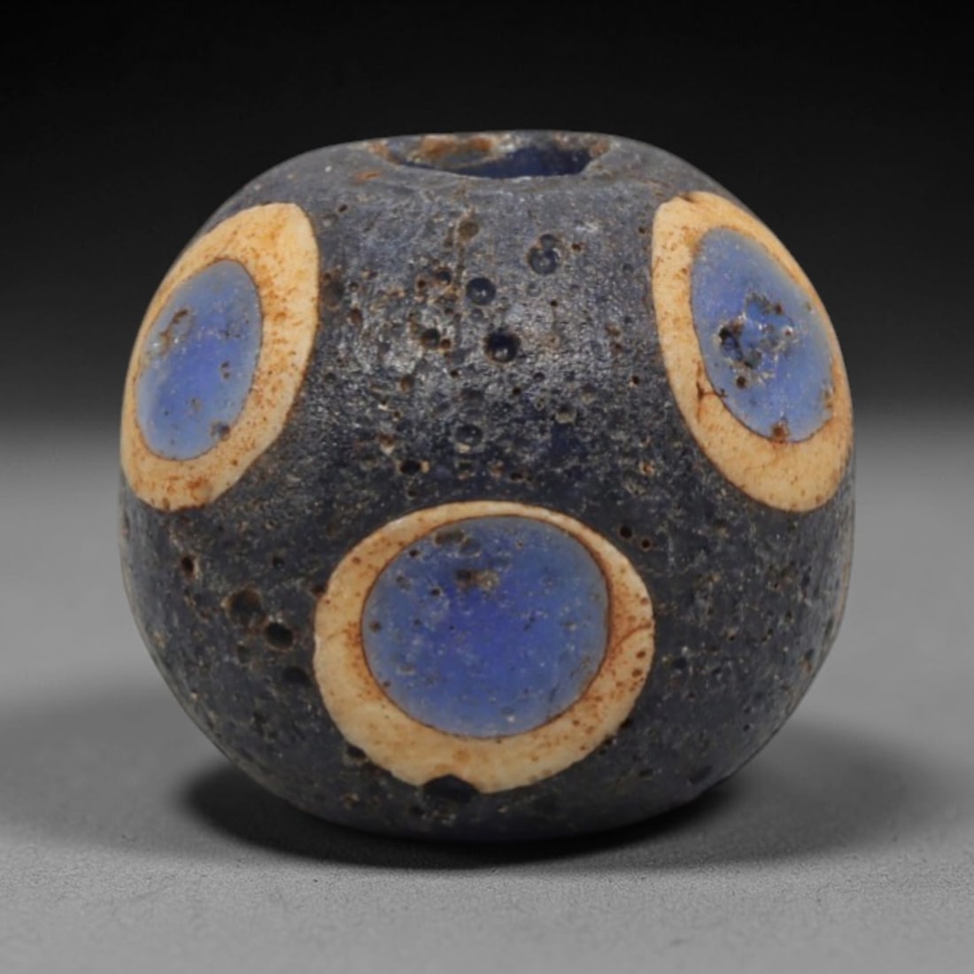 Ancient Glass Bead - Roman Egyptian Eye Amulet, 82ZN: Crafted from opaque glass with polychrome inlay, this spherical bead features a central suspension hole and is decorated with repeating concentric eye motifs in cream and vibrant blue glass against a