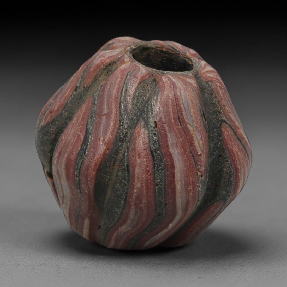 Ancient Islamic Ribbed Swirl Mosaic Glass Bead, 72ZN: A distinctive ancient Islamic mosaic glass bead, dating to approximately the 8th-12th centuries CE. This faceted, pumpkin-shaped bead features a dynamic ribbed swirl pattern of alternating deep pink/r
