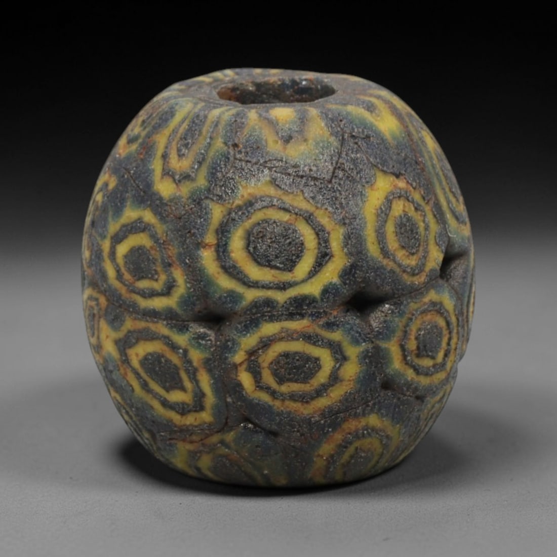Ancient Glass Bead - Roman Egyptian Eye Amulet, 55ZN: Crafted from opaque glass with polychrome inlay, this spherical bead features a central suspension hole and is decorated with repeating concentric eye motifs in yellow and dark grey glass against a da