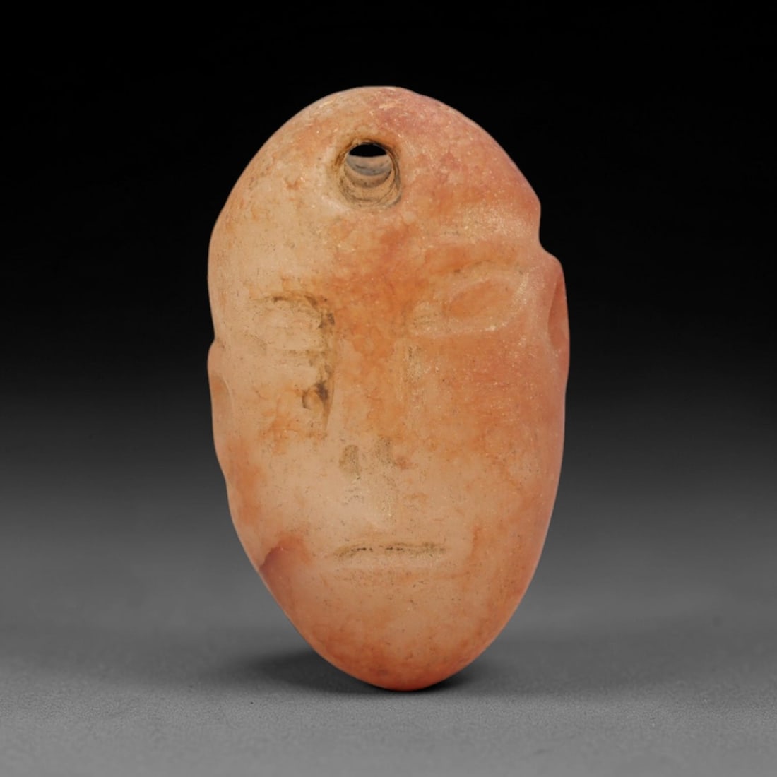 Ancient Carnelian Head Amulet - Levantine Figurine, 69XS: Carved from warm, translucent orange-red carnelian with natural tonal gradients and subtle surface patination, this amulet is formed as a stylized human head with incised linear detailing to define fa