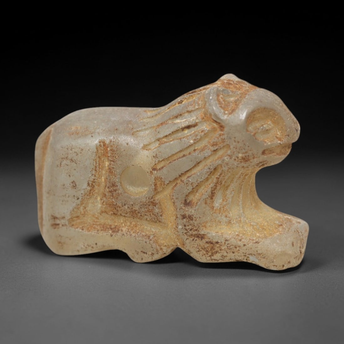 Ancient Glass Feline Amulet - Near Eastern Figurine, 20XV: Carved from translucent pale glass, this amulet depicts a stylized feline in a crouched posture, with incised linear striations defining the mane and simplified facial features. A circular perforation