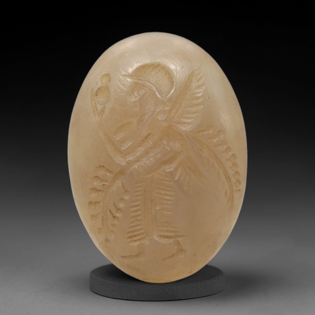 Ancient Carnelian Intaglio - Winged Deity Seal, 35XV: Crafted from polished carnelian, this oval intaglio seal depicts a winged deity in profile, adorned with a crested helmet and feathered wings. The figure holds a spherical object aloft in one hand, wh