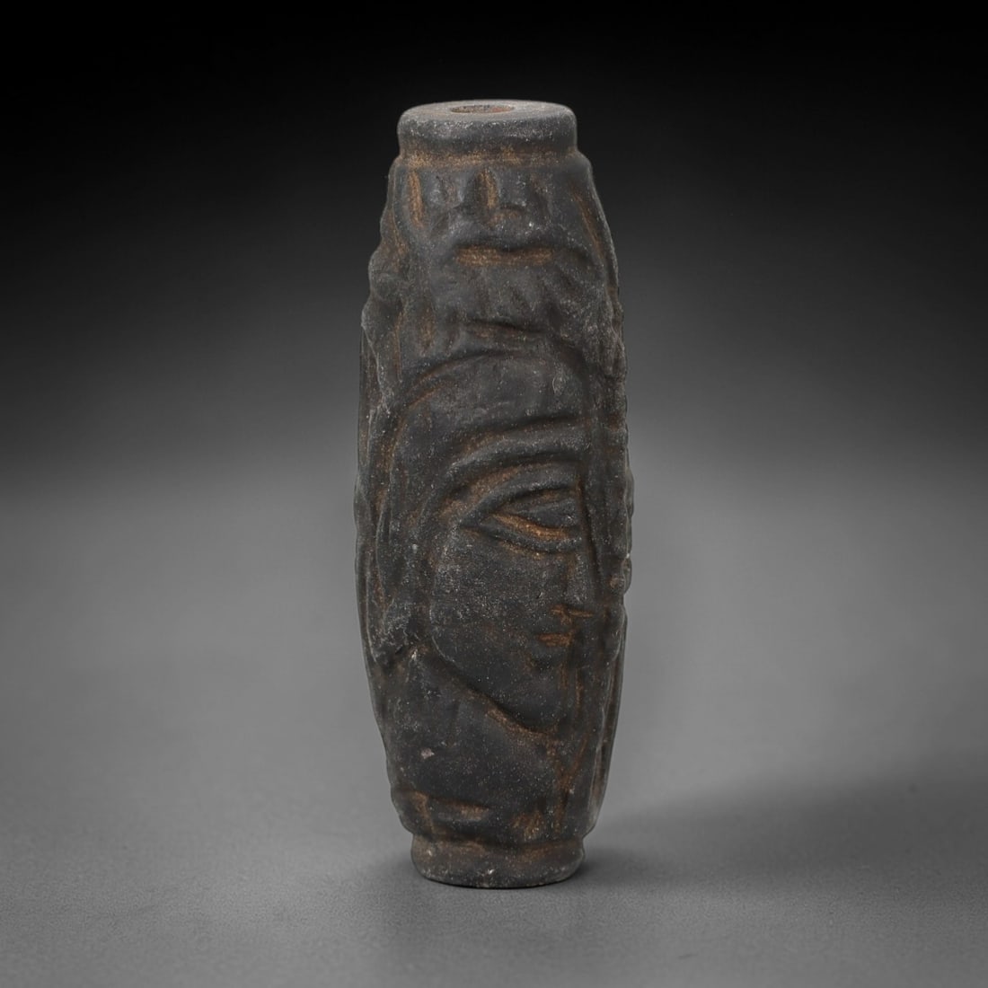 A Near Eastern Stone Cylinder Bead, Circa 1st Millennium BCE, 17FT: Carved from a dark, granular stone with a weathered patina, the object is a cylindrical bead with a central perforation for suspension. The exterior is decorated with a continuous carved relief depict