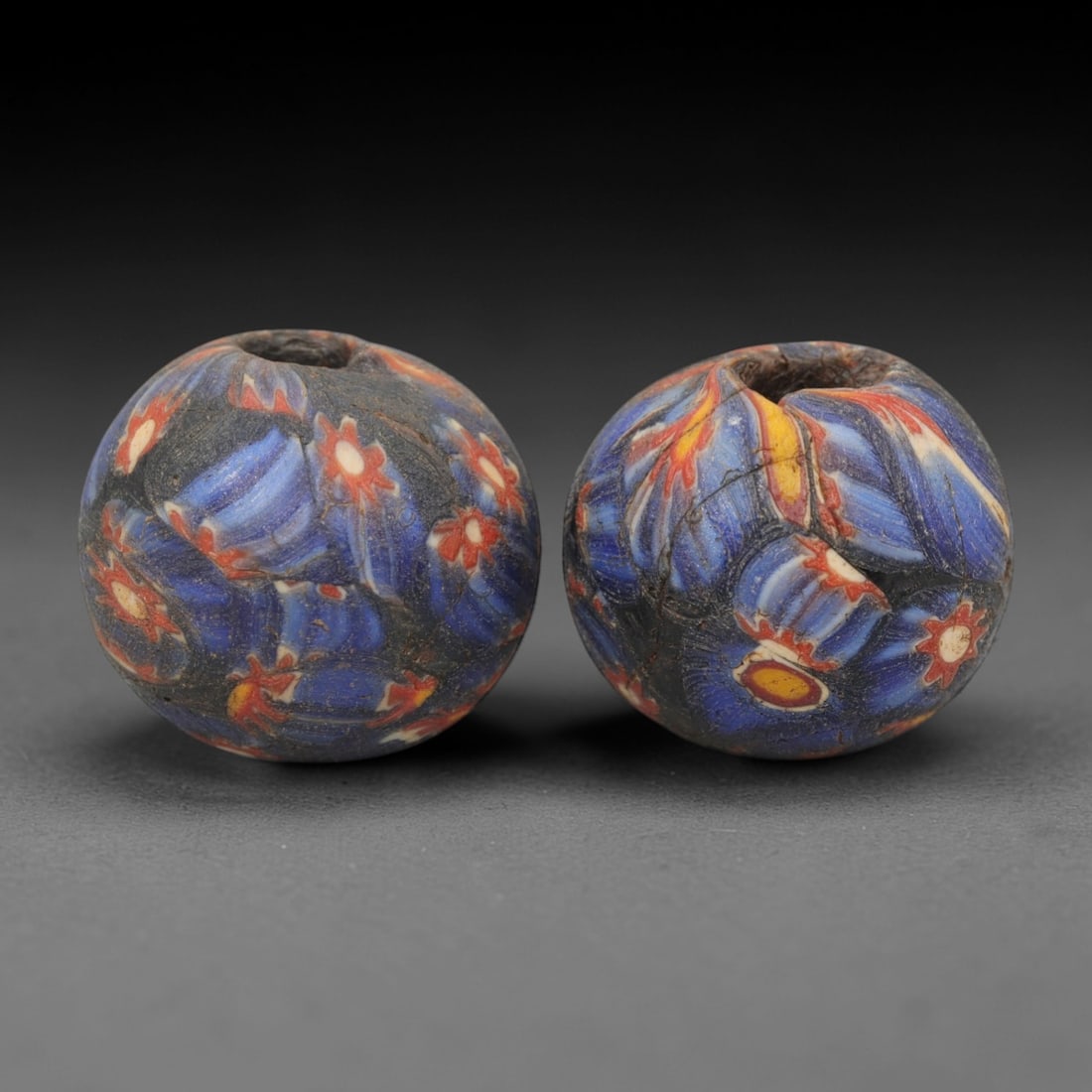 A Pair of Venetian Millefiori Glass Beads, 19th-20th Century, 42AD: Fabricated from opaque dark blue glass, each bead is formed as a sphere with a central drilled perforation. The surface is decorated with characteristic millefiori canes, featuring floral and leaf-lik