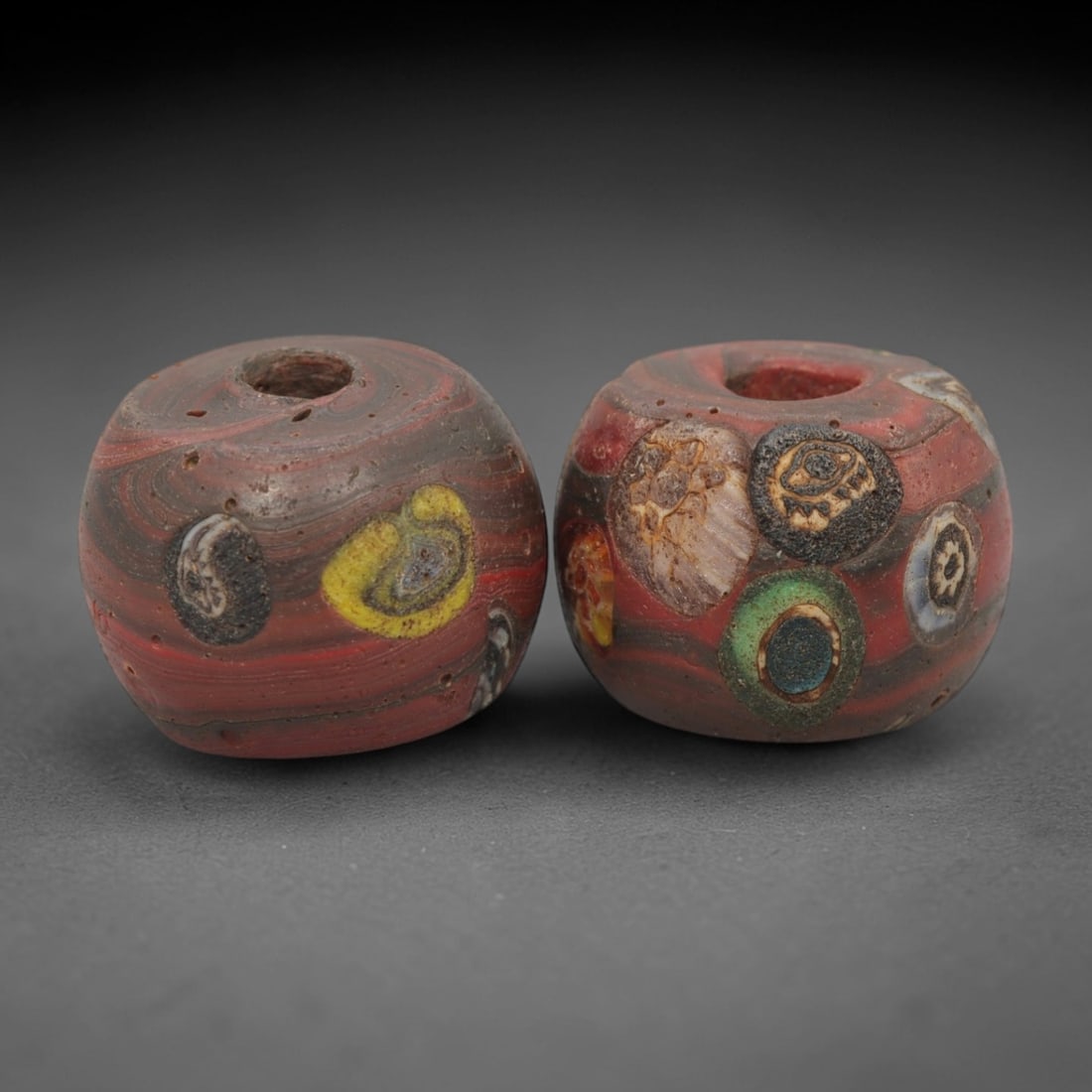 A Pair of Venetian Millefiori and Marbled Glass Beads, 19th-20th Century, 79AD: Fabricated from opaque red glass, each bead is formed as a short cylinder with a central drilled perforation. The surface combines marbled swirled patterns in red, brown, and black with characteristic