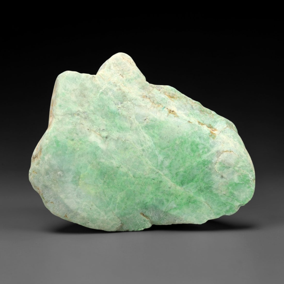 A Burmese Jadeite Jade Rough, 20th Century, 52FS: A piece of Burmese jadeite jade rough, with a pale greenish-white matrix and vibrant green mineral inclusions. The rough is shaped as an irregular, flattened form, with visible natural fractures and a