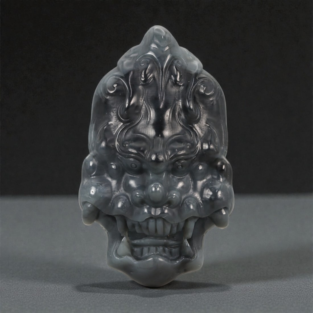 A Chinese Jade Mythical Beast Mask Carving, 20th Century, 63YA: Carved from dark gray to charcoal-toned jade with areas of translucency and subtle internal clouding, the object is modeled as a frontal mythical beast mask rendered in high relief. The face is symmet
