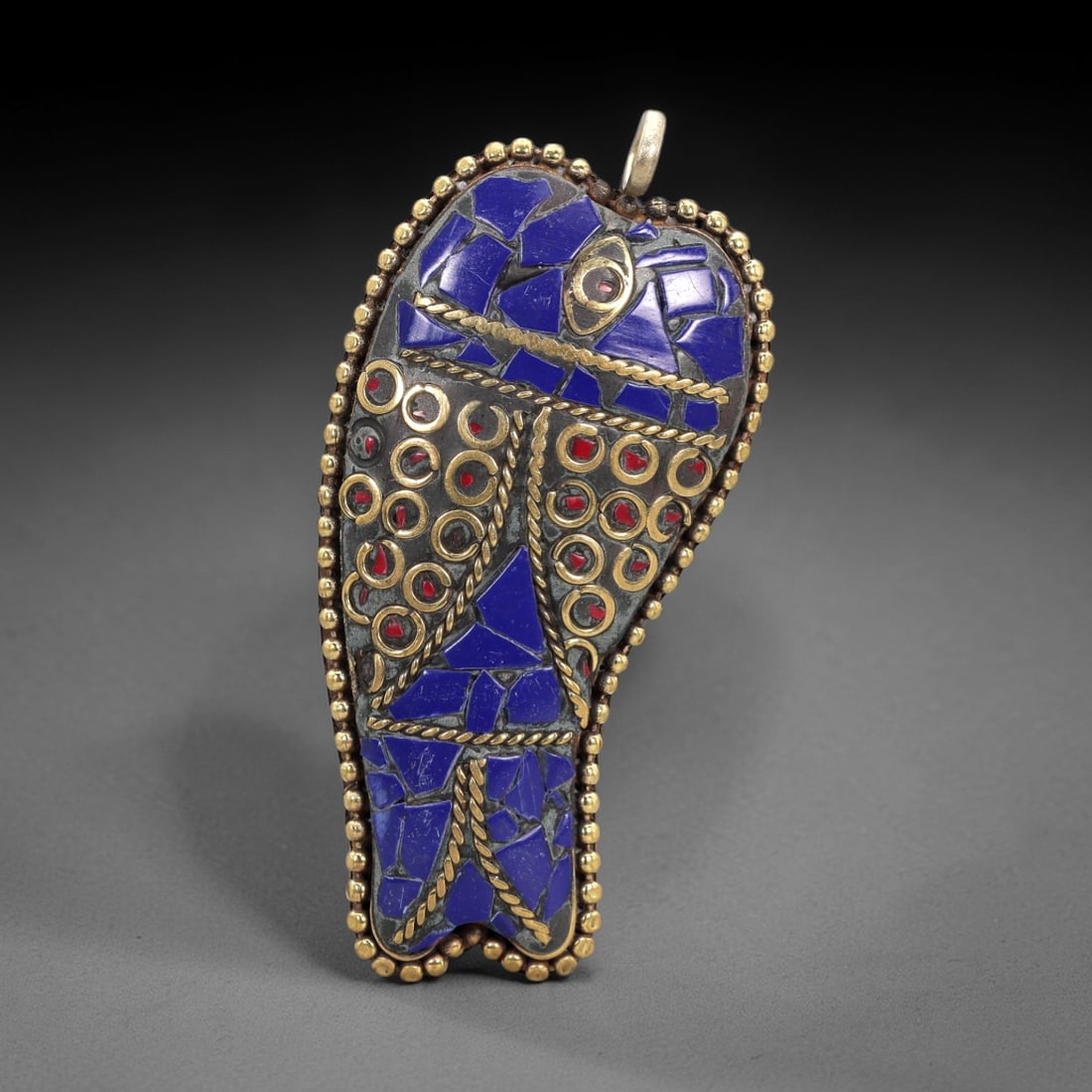 A Tibetan Silver Pendant with Lapis Lazuli and Red Stone Inlay, 20th Century, 98XM: Crafted from silver with a dark patinated base, the pendant is shaped as a stylized foot, with a cylindrical brass suspension loop at the top. The surface is inlaid with irregular fragments of lapis l