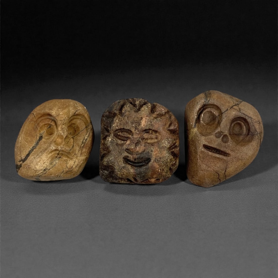 A Group of Three Medieval Stone Maskette Amulets, 10th-14th Century, 57FP: The group comprises three stone maskette amulets, each carved from distinct stone types with varied surface treatments. The first is a light brown stone mask with an irregular form, featuring recessed