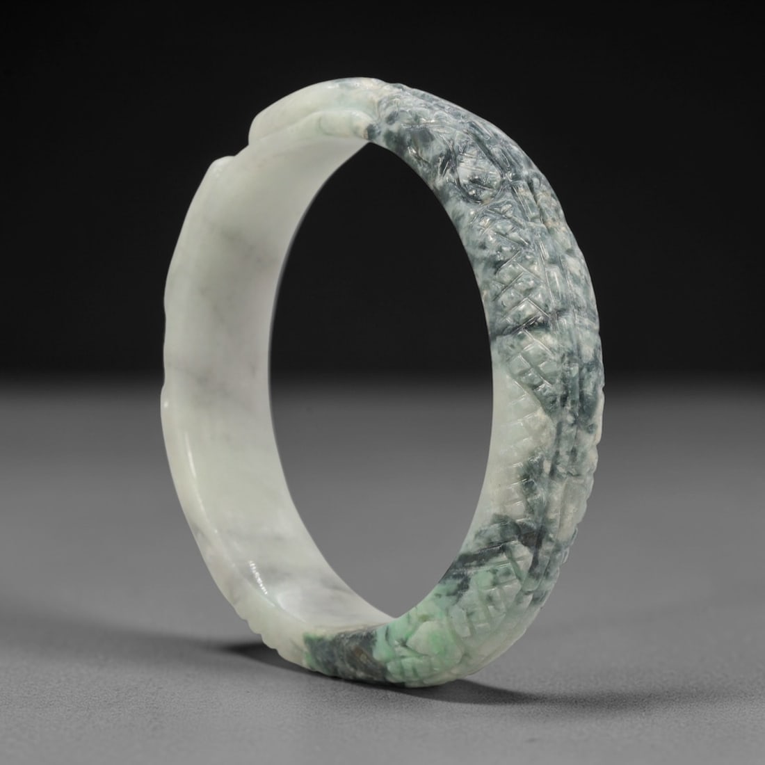A Chinese Carved Jadeite Jade Bangle Bracelet, 20th Century, 27ZL: A fine jadeite bangle of circular form, masterfully carved in relief with a continuous band of a sinuous dragon coiling around its circumference. The stone exhibits a striking variegated palette, with
