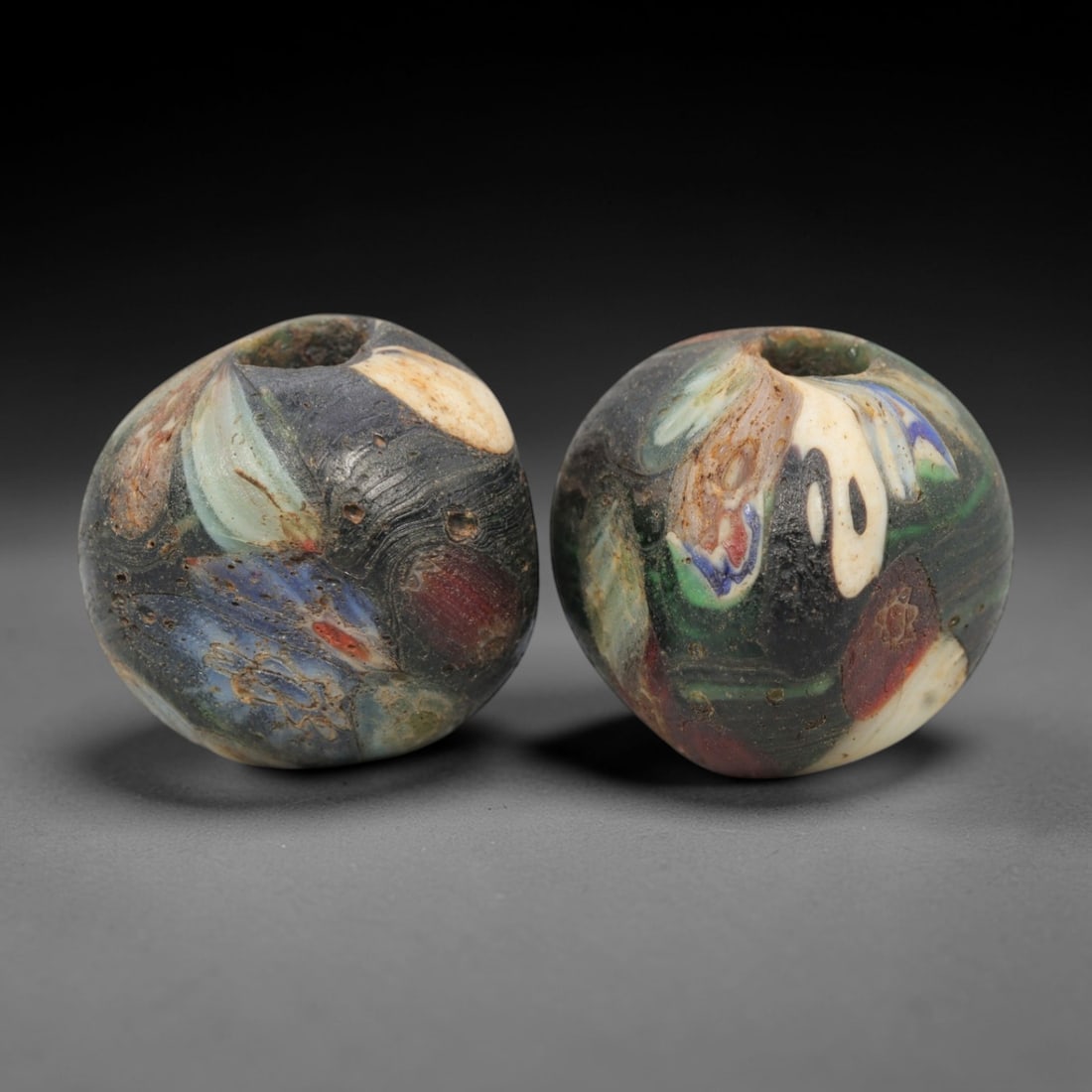 A Pair of Venetian Marbled Glass Beads, 19th-20th Century, 65AD: Fabricated from translucent and opaque glass, each bead is formed as a sphere with a central drilled perforation. The surface displays marbled patterns incorporating dark blue, green, white, red, and