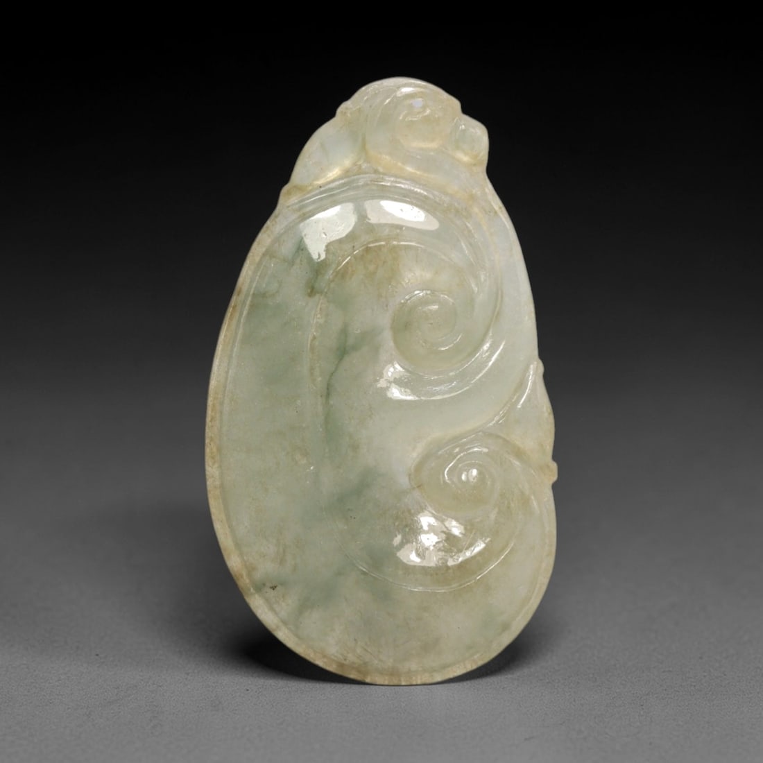 A Chinese Jadeite Jade Ruyi-Form Pendant, 20th Century, 62FR: Carved from translucent celadon jadeite jade with natural greyish inclusions and faint brownish tonal variations, the pendant is shaped in the form of a stylized ruyi scepter head. A simplified zoomor