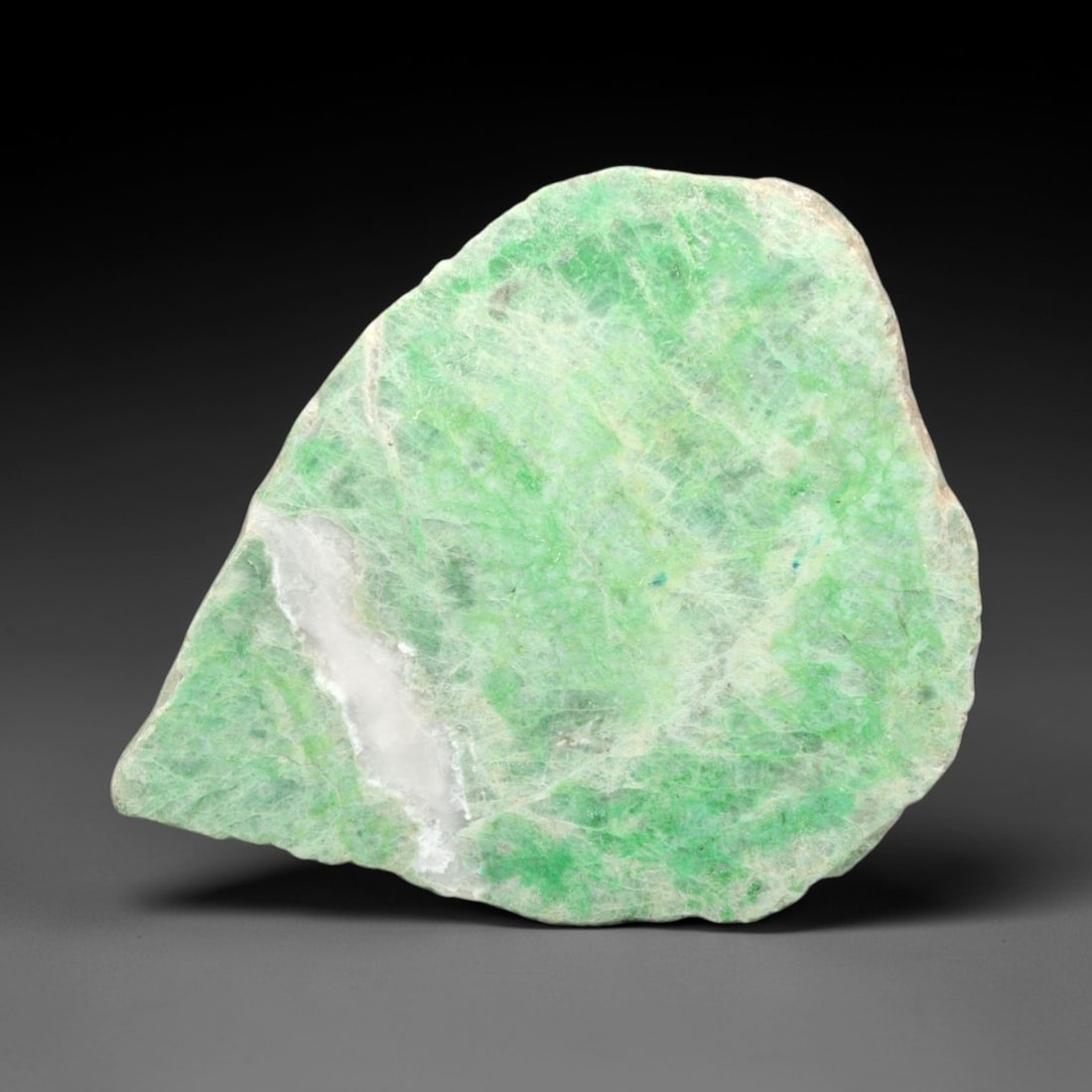 A Burmese Jadeite Jade Rough, 20th Century, 53FS: A piece of Burmese jadeite jade rough, with a pale greenish-white matrix and vibrant green mineral inclusions. The rough is shaped as an irregular, flattened form, with visible natural fractures and a