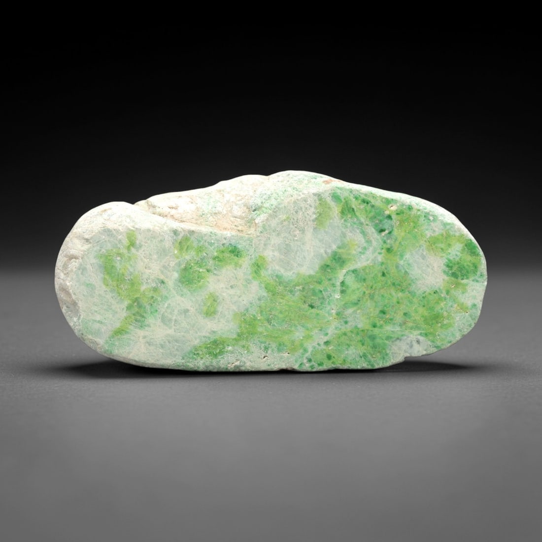 A Burmese Jadeite Jade Rough, 20th Century, 60FS: A piece of Burmese jadeite jade rough, with a pale greenish-white matrix and vibrant green mineral inclusions. The rough is shaped as an irregular, flattened, elongated form, with visible natural frac