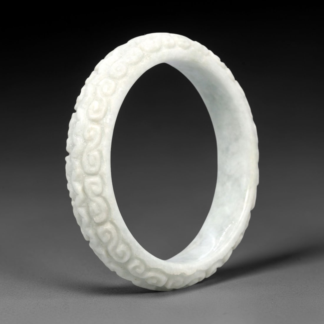 A Chinese Jadeite Jade Bangle Bracelet, 20th Century, 27FS: Carved from pale milky-white jadeite jade with a smooth, polished finish, the bangle is formed as a continuous circular band. The exterior surface is decorated with raised relief carving depicting con