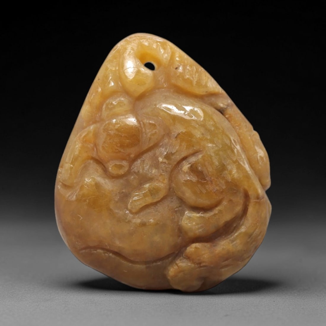 A Chinese Jadeite Jade Beast Pendant, 20th Century, 50FR: Carved from warm brownish-yellow jadeite jade with natural tonal variation and subtle mineral inclusions, the pendant is shaped as a teardrop form with a top suspension hole. A reclining beast is rend