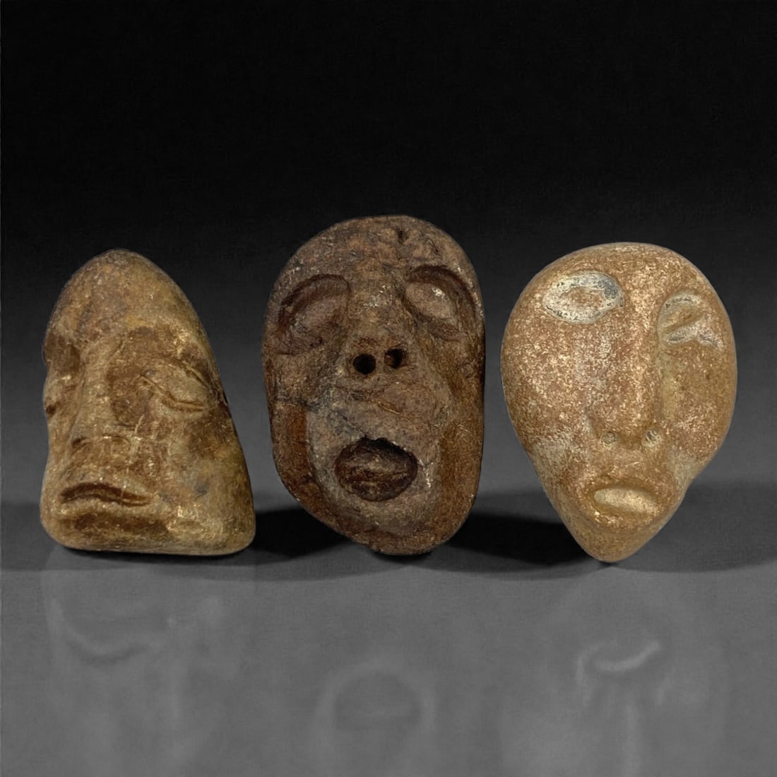 A Group of Three Medieval Stone Maskette Amulets, 10th-14th Century, 54FP: The group comprises three stone maskette amulets, each carved from distinct stone types with varied surface treatments. The first is a light brown stone mask with a teardrop form, featuring incised li