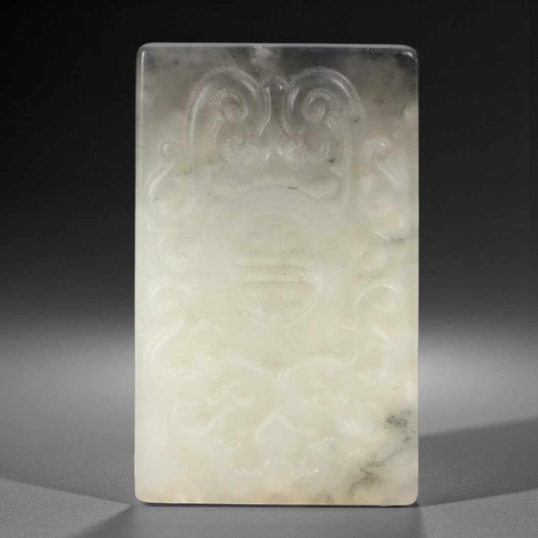 A Chinese Hetian Jade Plaque, 20th Century, 79FH: Carved from translucent Hetian jade with subtle grey inclusions, the rectangular plaque features low-relief decoration of symmetrical scrollwork flanking a central shou (longevity) symbol. The upper s