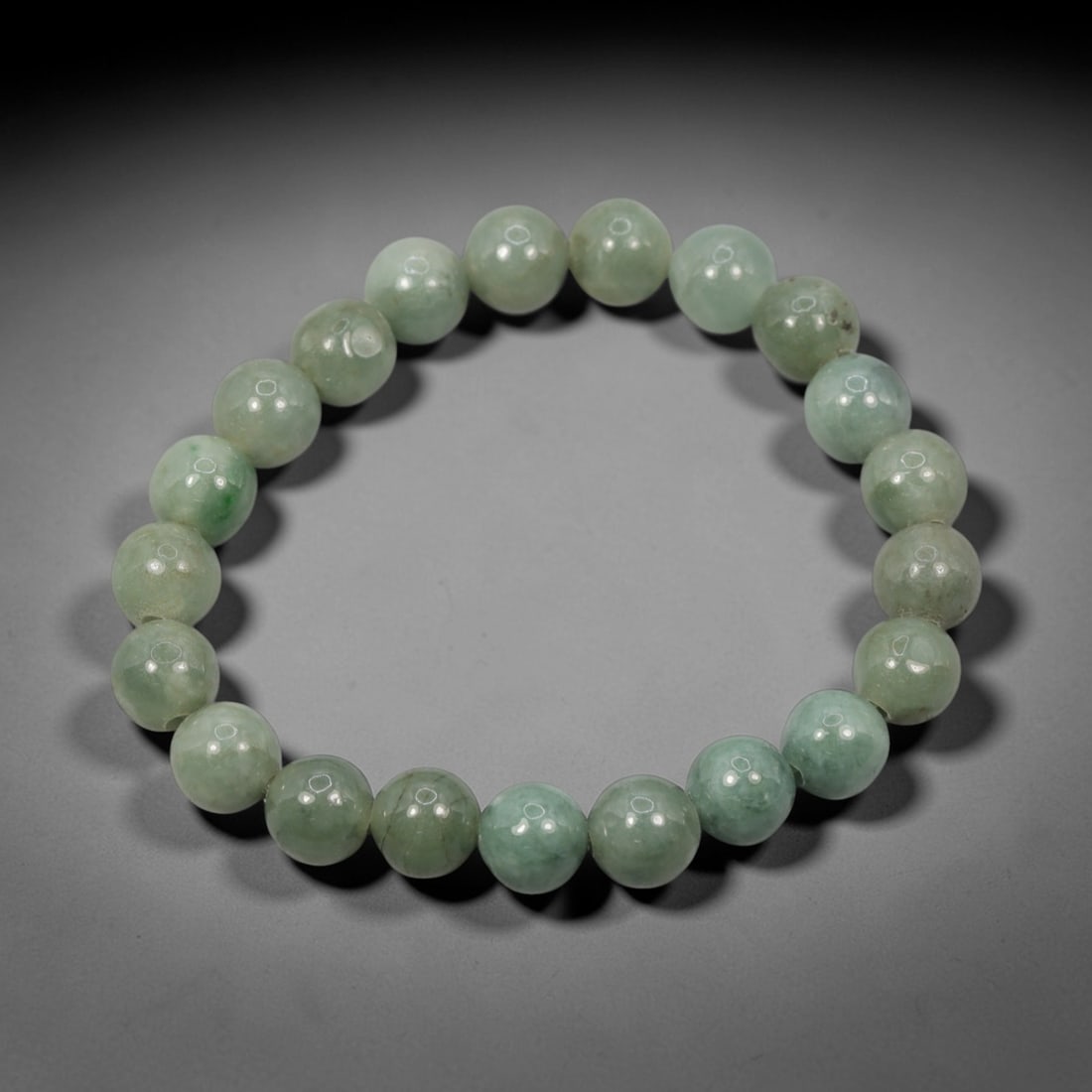 A Chinese Jadeite Jade Bead Bracelet, 20th Century, 55ZK: Composed of a single strand of spherical jadeite jade beads forming a circular bracelet. The beads display natural tonal variation from pale green to light green, with visible internal mineral inclusi