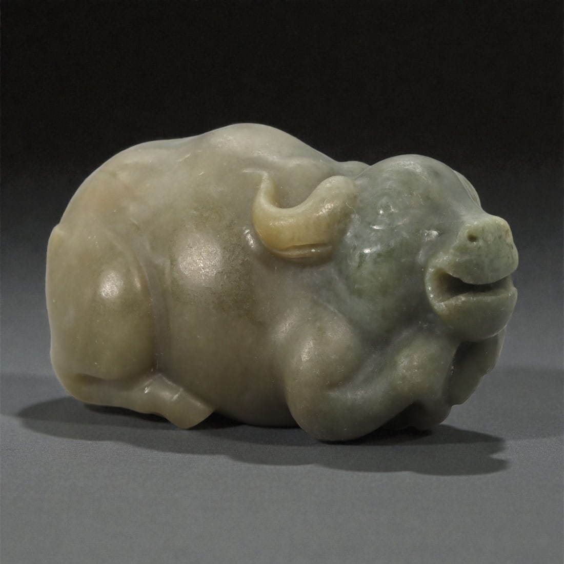A Chinese Nephrite Jade Buffalo Figure, 20th Century, 97YA: Carved from pale green to buff-toned nephrite jade with areas of soft translucency and natural tonal variation, the figure represents a recumbent buffalo shown in a compact, resting posture. The head