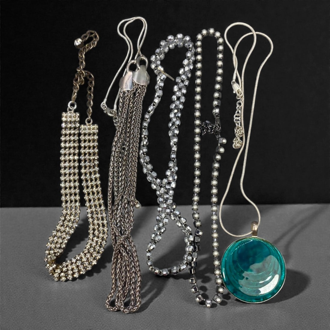 An Estate Lot of Five Necklaces, Late 20th Century, 59FK: The lot comprises five necklaces constructed from mixed materials, including silver-toned and gunmetal-toned metal chains, rhinestones, and resin. One piece features a double-strand pave rhinestone de