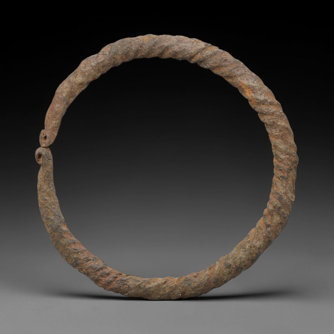 An Ancient Iron Twisted Bracelet, 1st-3rd Century CE, 27FV: Forged from iron with a continuous, spiraled twist along its entire length, the bracelet is formed as a near-complete circle with a narrow terminal gap. The terminals are shaped into small, interlocki