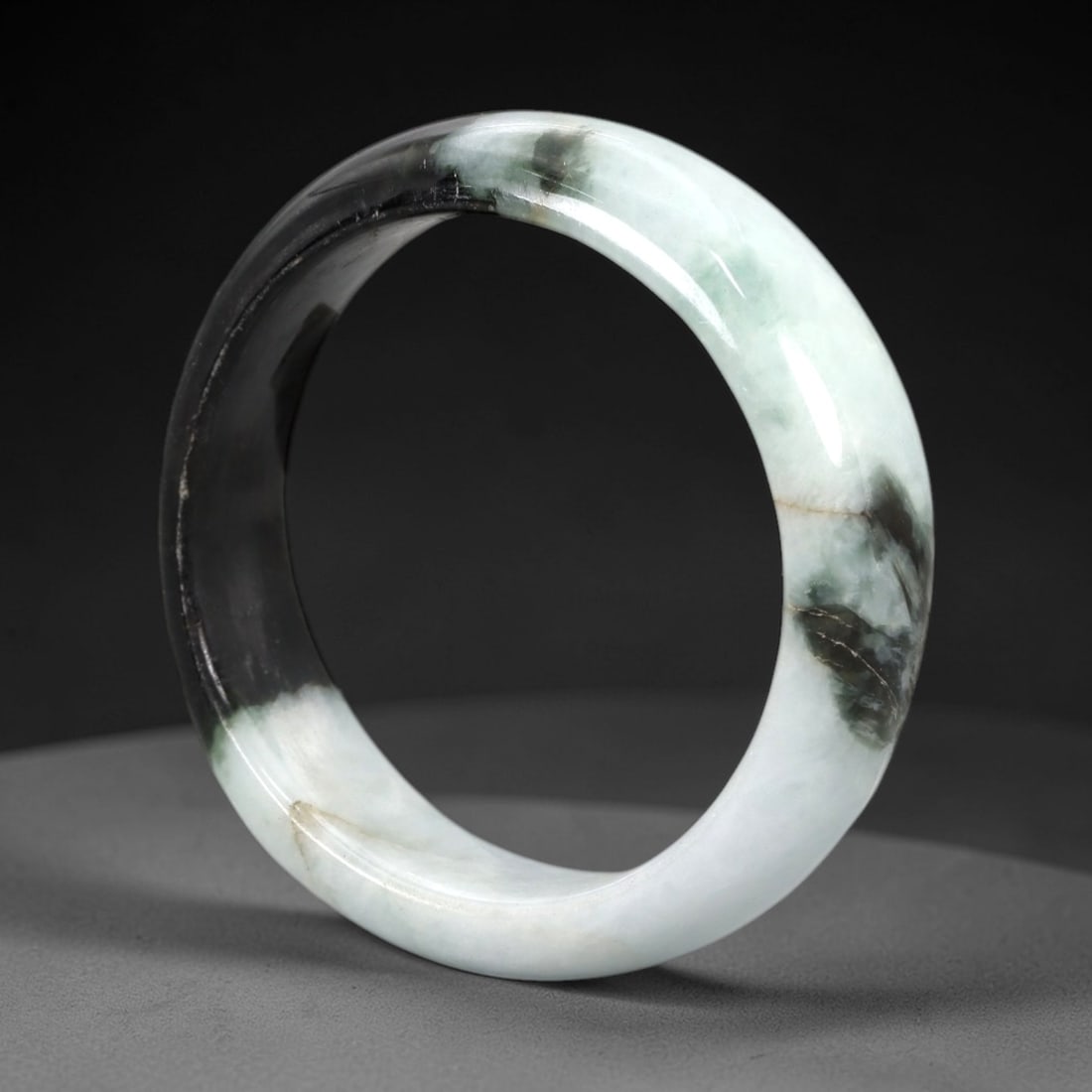 A Chinese Jadeite Jade Bangle Bracelet, 20th Century, 99ZK: Carved from jadeite jade with a striking bi-color composition of opaque pale white and translucent deep blackish-green, the bangle is formed as a solid circular band with a flattened rounded cross-sec