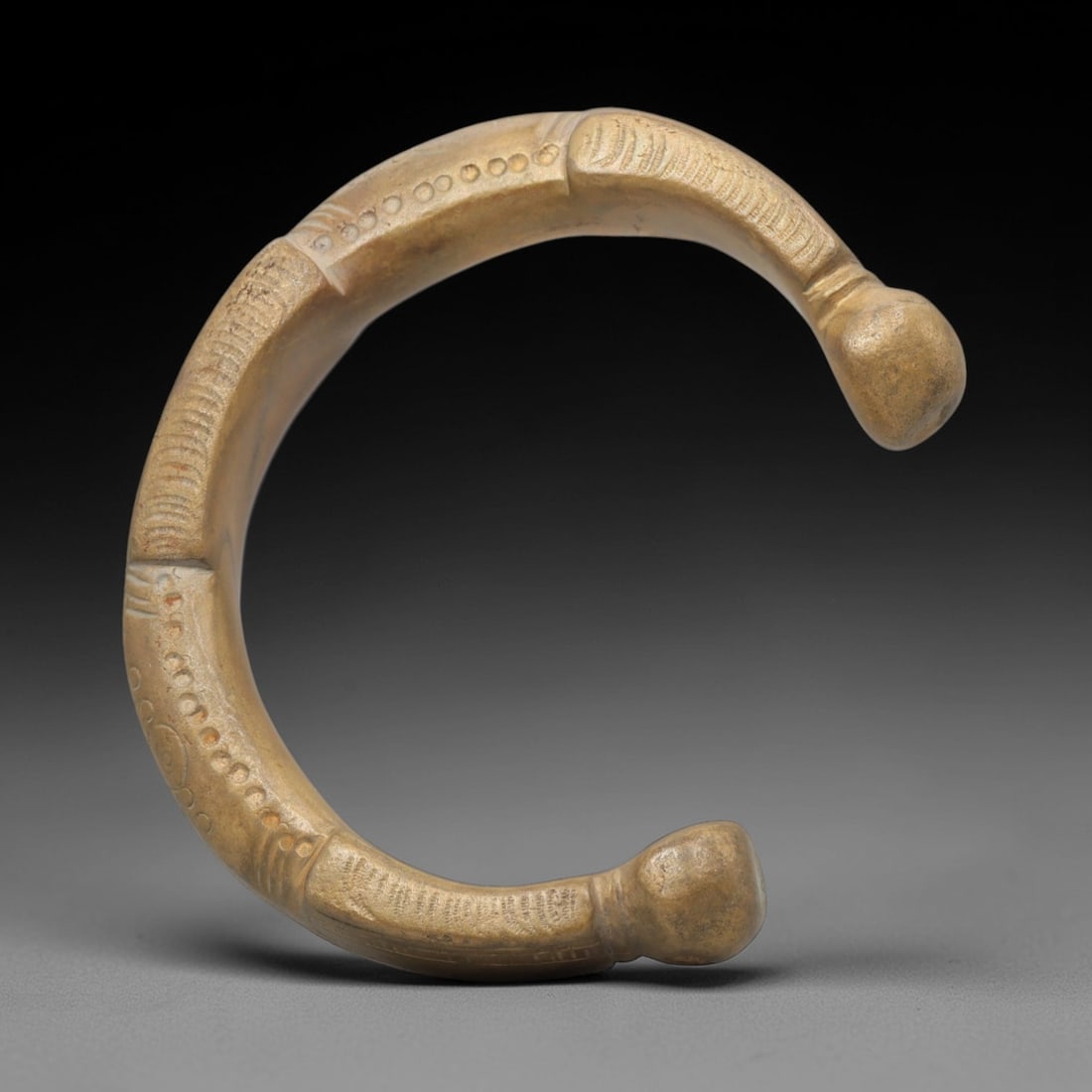 An African Bronze Manila Bracelet, 19th Century, 89FT: Cast from bronze with a flattened, slightly curved section, the bracelet is formed as an open C-shape with rounded, bulbous terminals. The surface is decorated with incised linear striations and a con