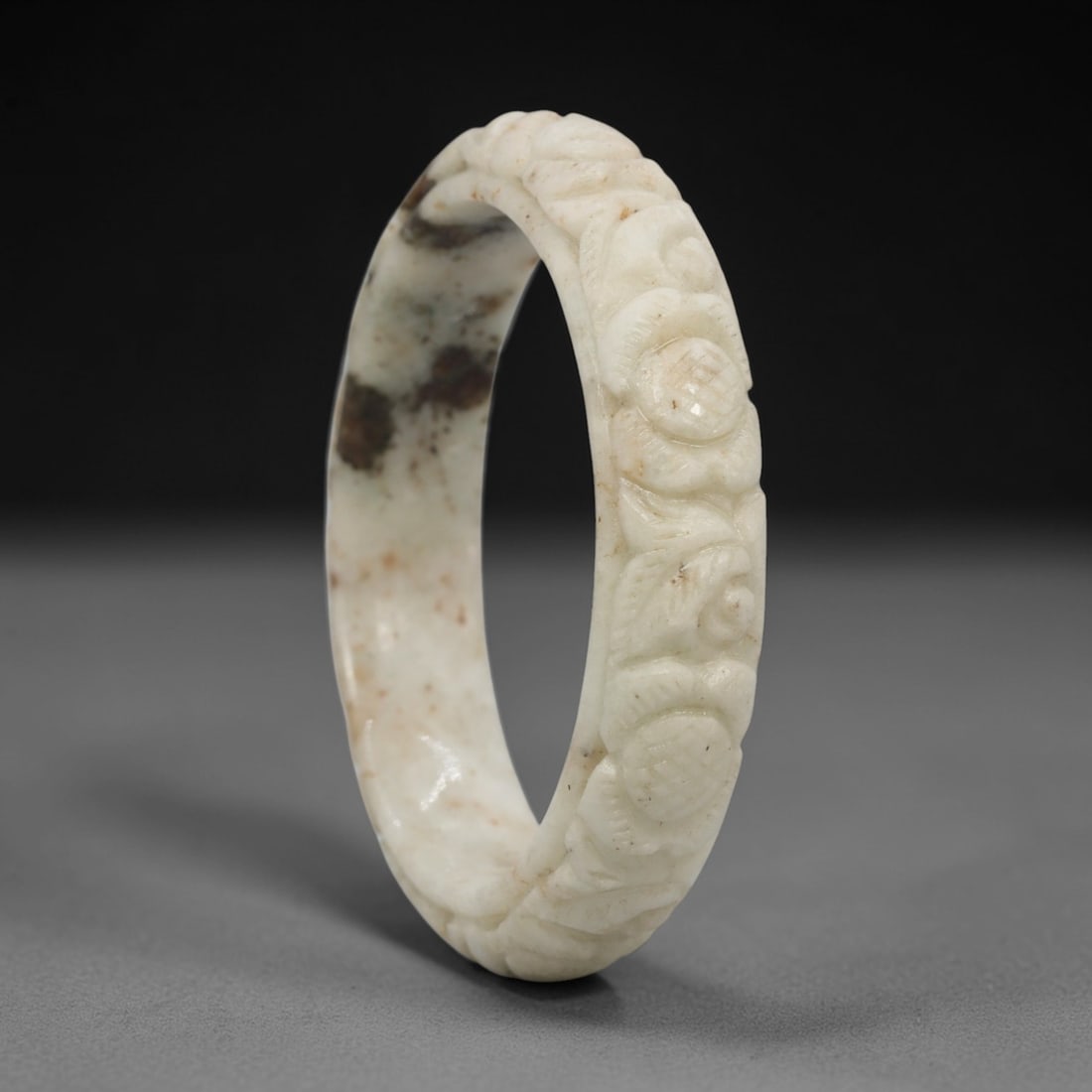 A Chinese Carved Jadeite Jade Bangle Bracelet, 20th Century, 20ZL: A fine jadeite bangle of flattened circular form, masterfully carved in relief with a continuous band of floral blossoms and scrolling foliate motifs. The stone exhibits a creamy white base, heavily a