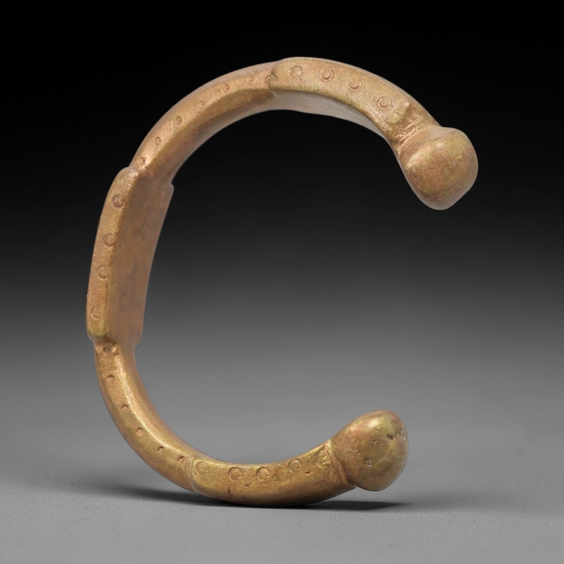 An African Bronze Manila Bracelet, 19th Century, 94FT: Cast from bronze with a flattened, slightly curved section, the bracelet is formed as an open C-shape with rounded, bulbous terminals. The surface is decorated with evenly spaced, incised circular mot