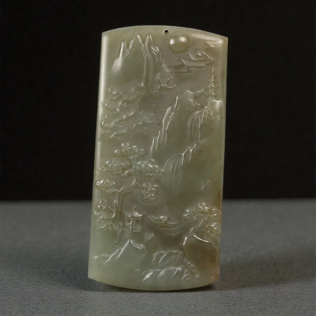 A Chinese Nephrite Jade Landscape Plaque, 20th Century, 81FC: Carved from pale celadon to grey-green nephrite jade with subtle tonal variation and minor natural inclusions, the object is worked as a vertically oriented plaque with a gently curved upper edge. The