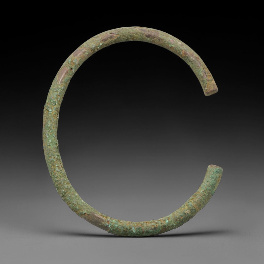 A Roman Bronze Bracelet, 1st-3rd Century CE, 26FV: Cast from bronze with a thick, rounded section, the bracelet is formed as an open C-shape with rounded, slightly tapered terminals. The surface is covered in a patchy green patina with extensive reddi