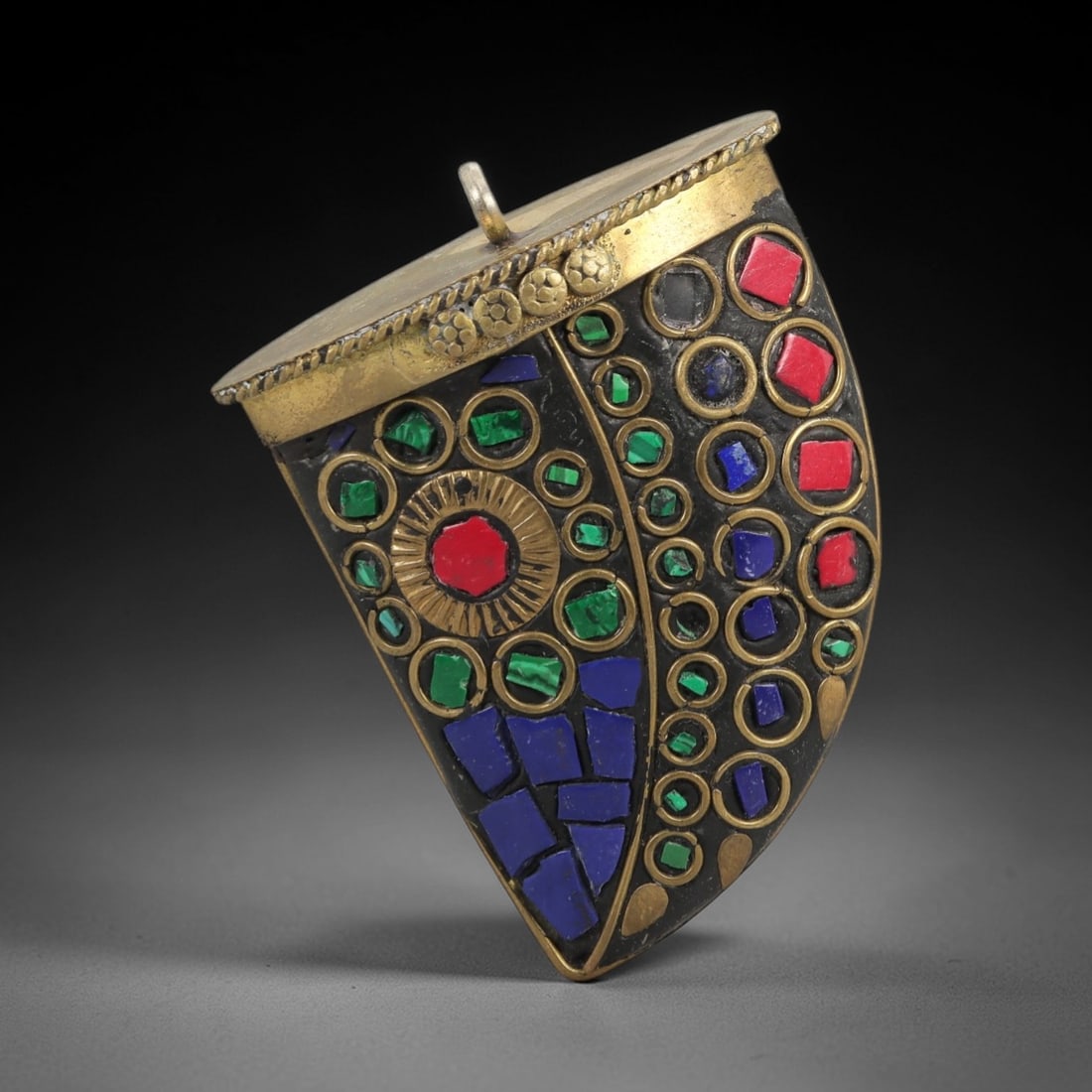 A Tibetan Gilt-Metal and Mosaic Gau Pendant, 20th Century, 86ZL: This Tibetan gau (prayer amulet) is of distinctive shield-shaped form, crafted from a gilt-brass alloy with a dark, aged patina. The surface is adorned with a vibrant mosaic of colored composite stone