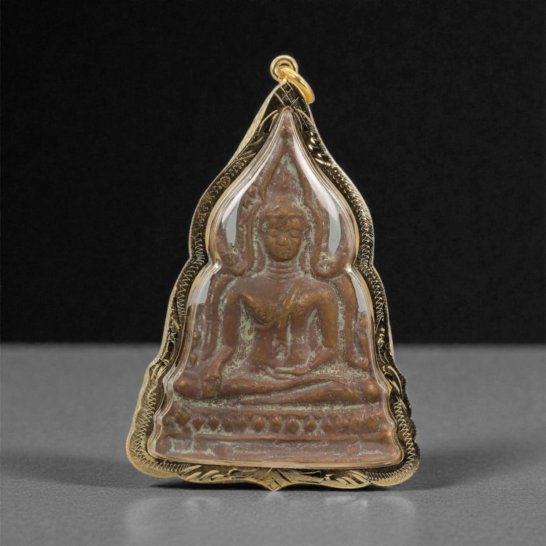 A Thai Copper Buddha Amulet in Gilt Metal Frame, 20th Century, 49FN: The amulet is encased in a stylized, pointed-arch gilt metal frame with incised decorative borders and a suspension loop at the apex. The obverse features a copper relief of a seated Buddha in bhumisp