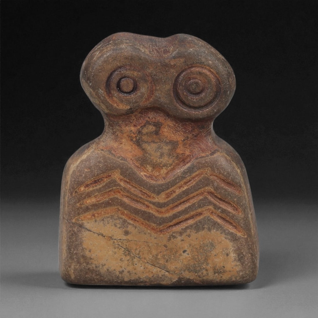An Ancient Near Eastern Stone Eye Idol, 4th-3rd Millennium BCE, 19AB: Carved from a single piece of stone with a natural earthy brown and beige patina, the idol features a stylized head with two prominent, concentric circular eyes, each defined by incised lines. The hea