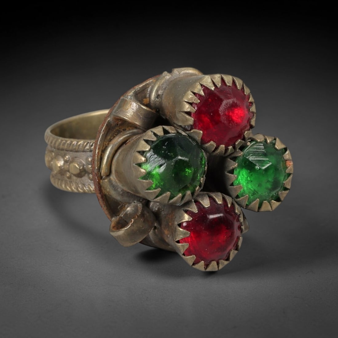 A Turkish Silvered Metal and Colored Paste Ring, 20th Century, 73AC: Crafted from a silvered metal alloy with a warm, aged patina, the ring features a wide, flat band decorated with raised beaded motifs and twisted ropework detailing along the edges. The bezel is set w