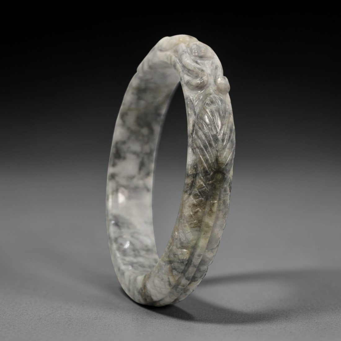 A Chinese Carved Jadeite Jade Bangle Bracelet, 20th Century, 78ZL: A fine jadeite bangle of circular form, masterfully carved in relief with a continuous band of a sinuous dragon coiling around its circumference. The stone exhibits a striking variegated palette, with