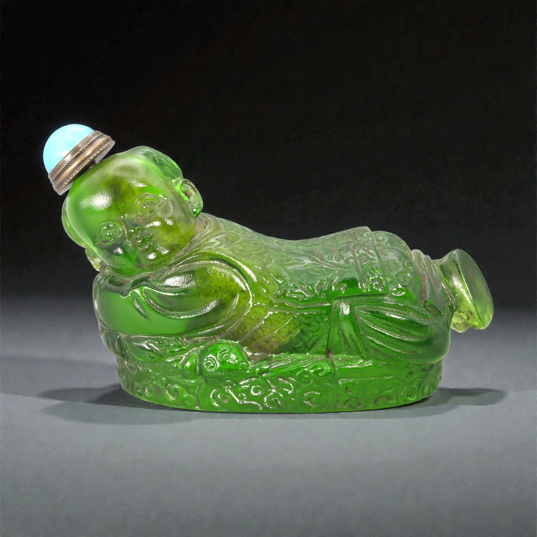 A Chinese Glass Figural Snuff Bottle, Qing Dynasty, Late 19th-Early 20th Century, 39FC: Molded from translucent green glass, the bottle is formed as a reclining child figure resting on one side atop an integrated base. The head is turned slightly toward the viewer, with softly modeled fa