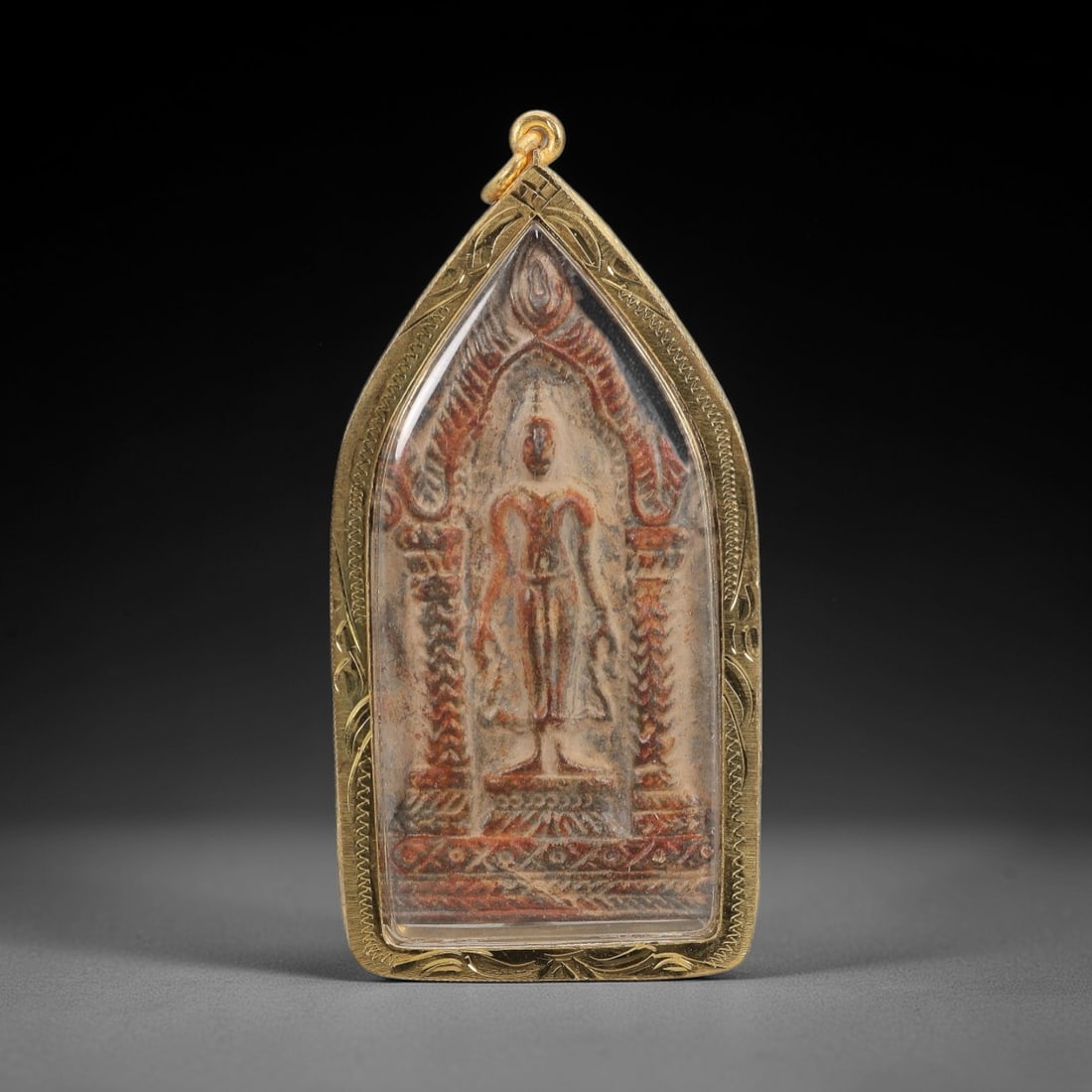 A Thai Leela Buddha Amulet in a Gilded Metal Frame, 20th Century, 80FT: The amulet is a rectangular plaque with a pointed upper edge, depicting the Buddha in the Leela (walking) pose, flanked by ornate pillars and a flaming aureole. The plaque is composed of a sacred powd