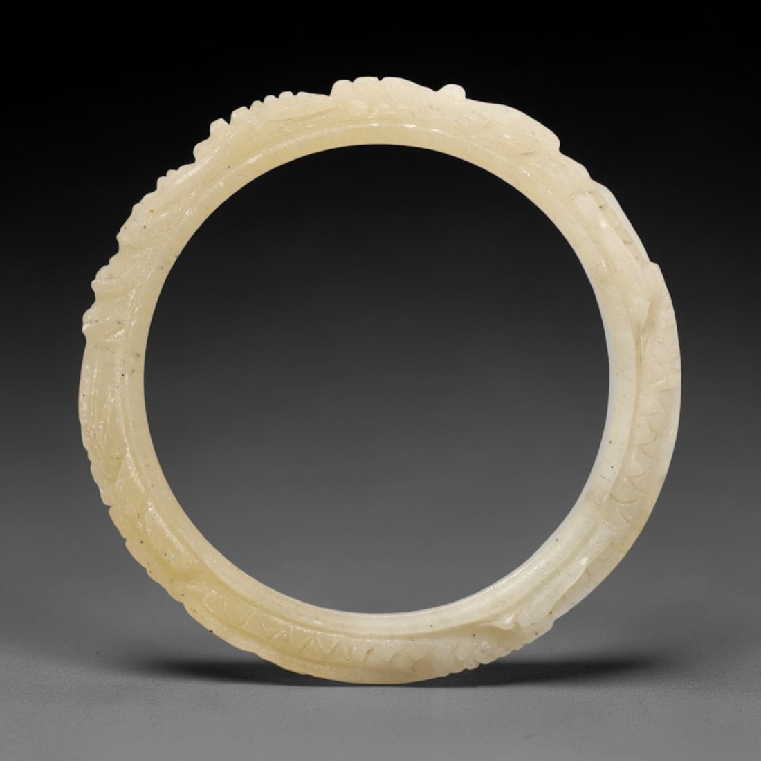 A Chinese Jadeite Jade Bangle Bracelet, 20th Century, 39FS: Carved from pale creamy-yellow jadeite jade with fine dark inclusions, the bangle is formed as a continuous circular band. The exterior surface is decorated with raised relief carving depicting a coil