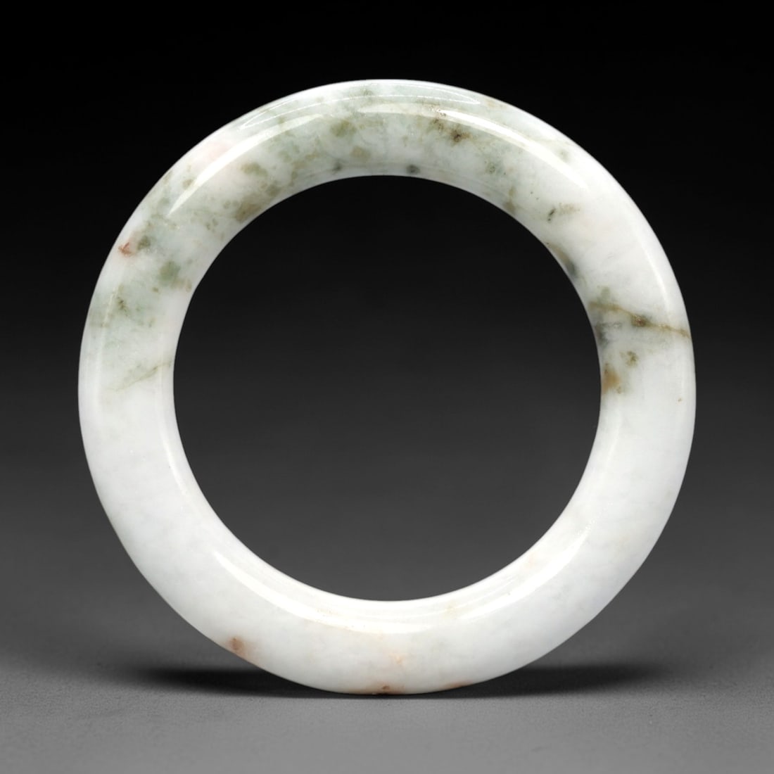 A Burmese Jadeite Jade Bangle, 20th Century, 3FS: A Burmese jadeite jade bangle, with a pale white matrix and scattered light green and russet-brown mineral inclusions. The bangle is of circular form, with a smooth, polished surface revealing the int