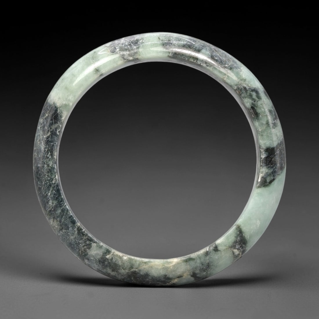 A Burmese Jadeite Jade Bangle, 20th Century, 21FS: A Burmese jadeite jade bangle, with a pale greenish-white matrix and prominent dark green mineral inclusions. The bangle is of circular form, with a smooth, polished surface revealing the internal cry
