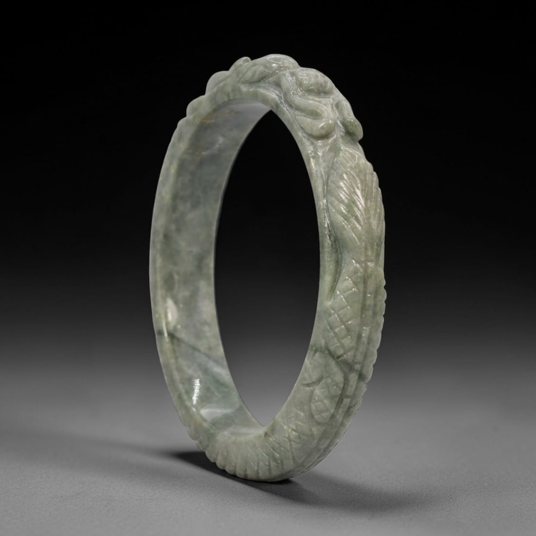 A Chinese Carved Jadeite Jade Bangle Bracelet, 20th Century, 25ZL: A fine jadeite bangle of circular form, masterfully carved in relief with a continuous band of a sinuous dragon coiling around its circumference. The stone exhibits a translucent pale green base, with